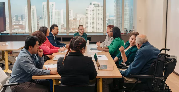 Several people sitting around a table with laptops and paper in front of them. A large window is behind them with tall buildings outside.