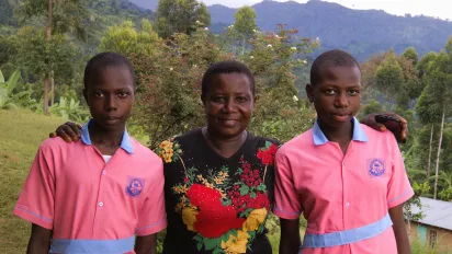 A portrait of Rebecca Nakayega Wandegge. She stands in a rural area, smiling, next to her two daughters who are wearing their school uniforms. (Uganda, 2025)