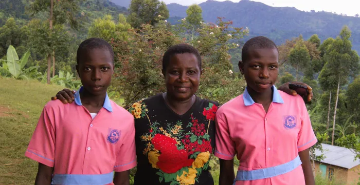 A portrait of Rebecca Nakayega Wandegge. She stands in a rural area, smiling, next to her two daughters who are wearing their school uniforms. (Uganda, 2025)